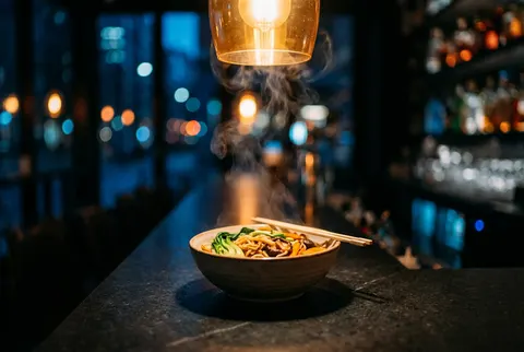 A steaming bowl of wok noodles on a dark stone counter under a warm pendant lamp late at night